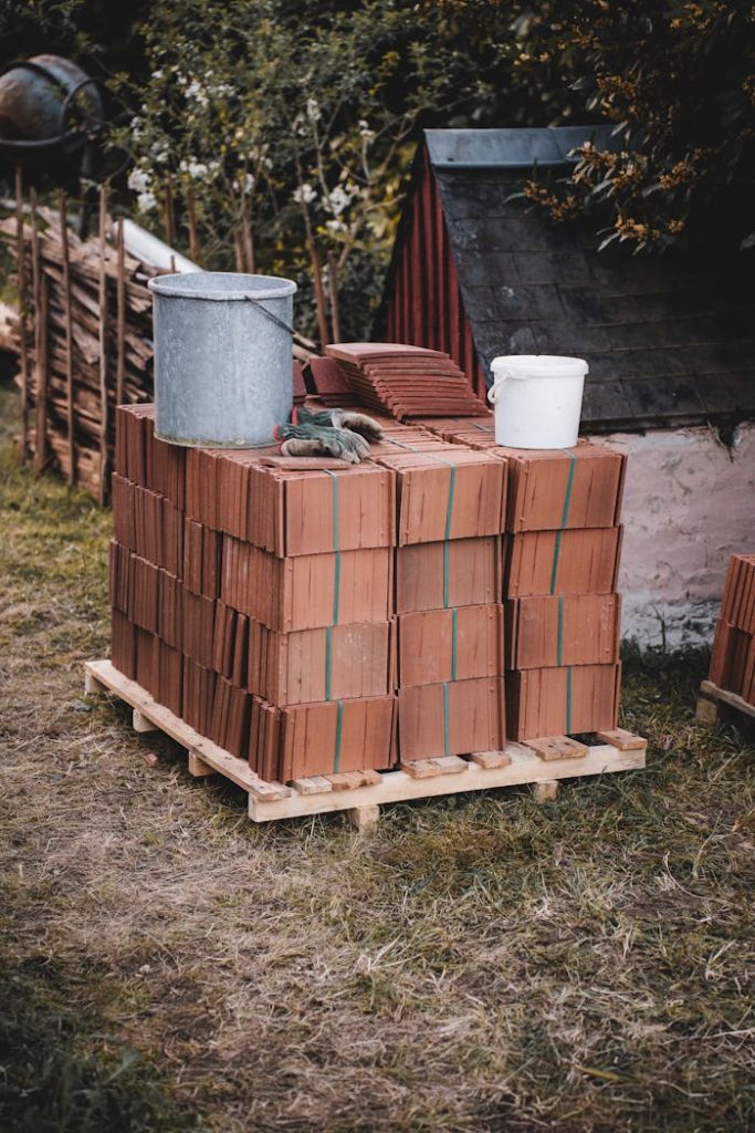 stack-of-roof-tiles-with-buckets-outdoors-31763542 Pallet of stacked roof tiles with metal and plastic buckets outside near a shed.